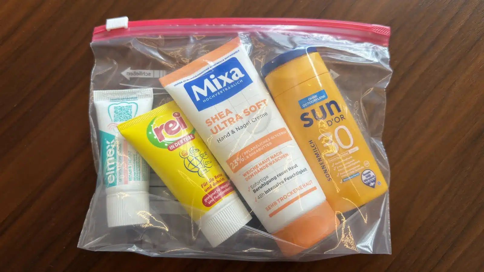 Durchsichtiger Beutel mit Zahnpasta, Reisewaschmittel, Handcreme und Sonnenmilch auf einem Holztisch