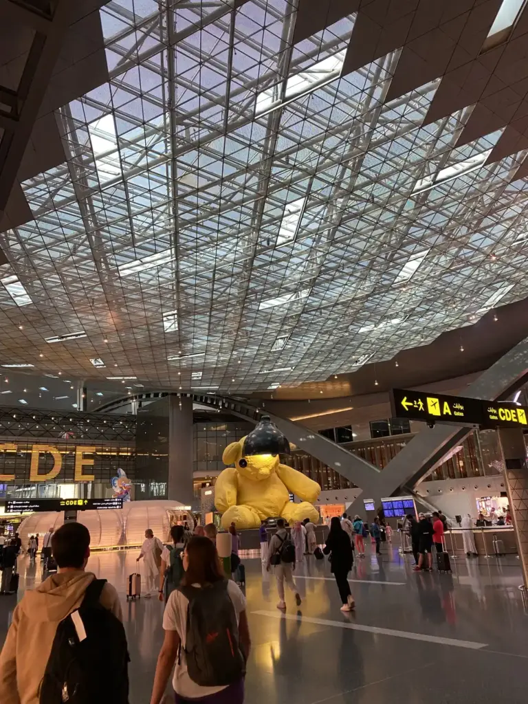 Gelber Bär mit Lampe auf der Duty Free Plaza des Flughafens Doha, darüber Wegweiser zu den Gates