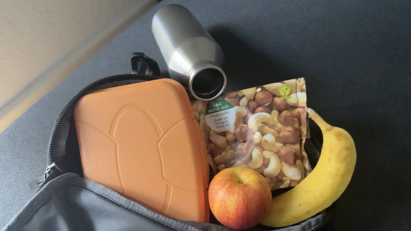 Handgepäck-Rucksack mit Brotdose, Nüssen, Apfel, Banane und offener, leerer Trinkflasche als Verpflegung für den Flug